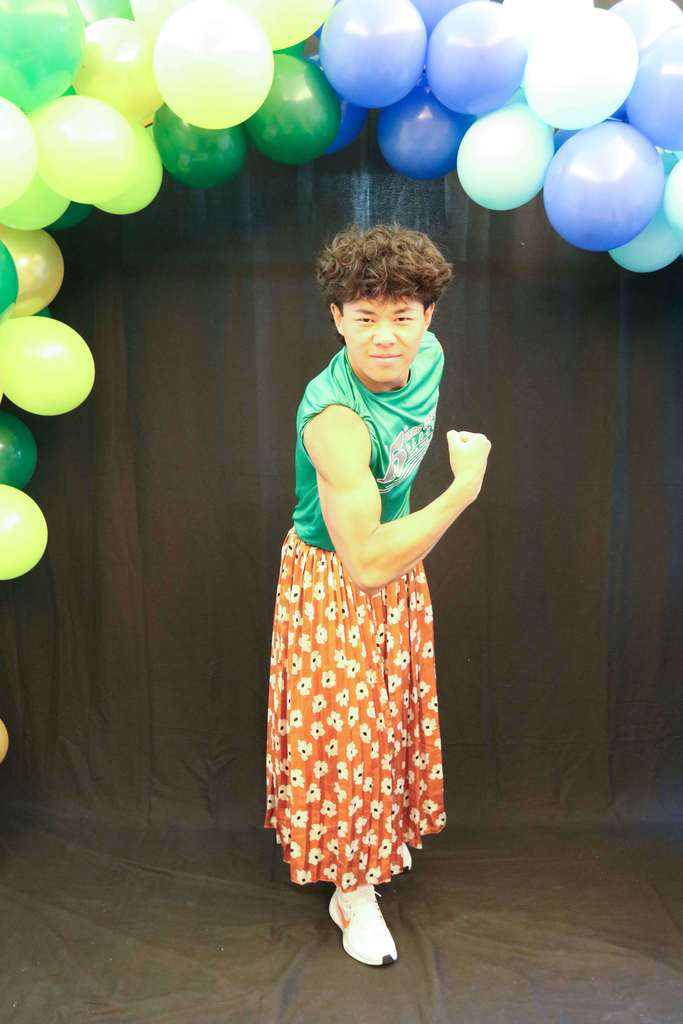 A student is wearing a green t-shirt with orange with flower skirt showing off his muscle arms for a photo. 