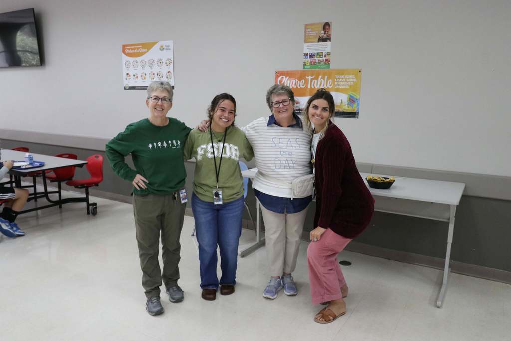 Four staff members are dressed like the elements colors smiling for a photo together inside the cafeteria. 