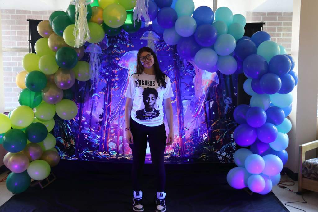 A student wearing her class color white shirt smiling for a photo with the blue light and balloon arch around her. 