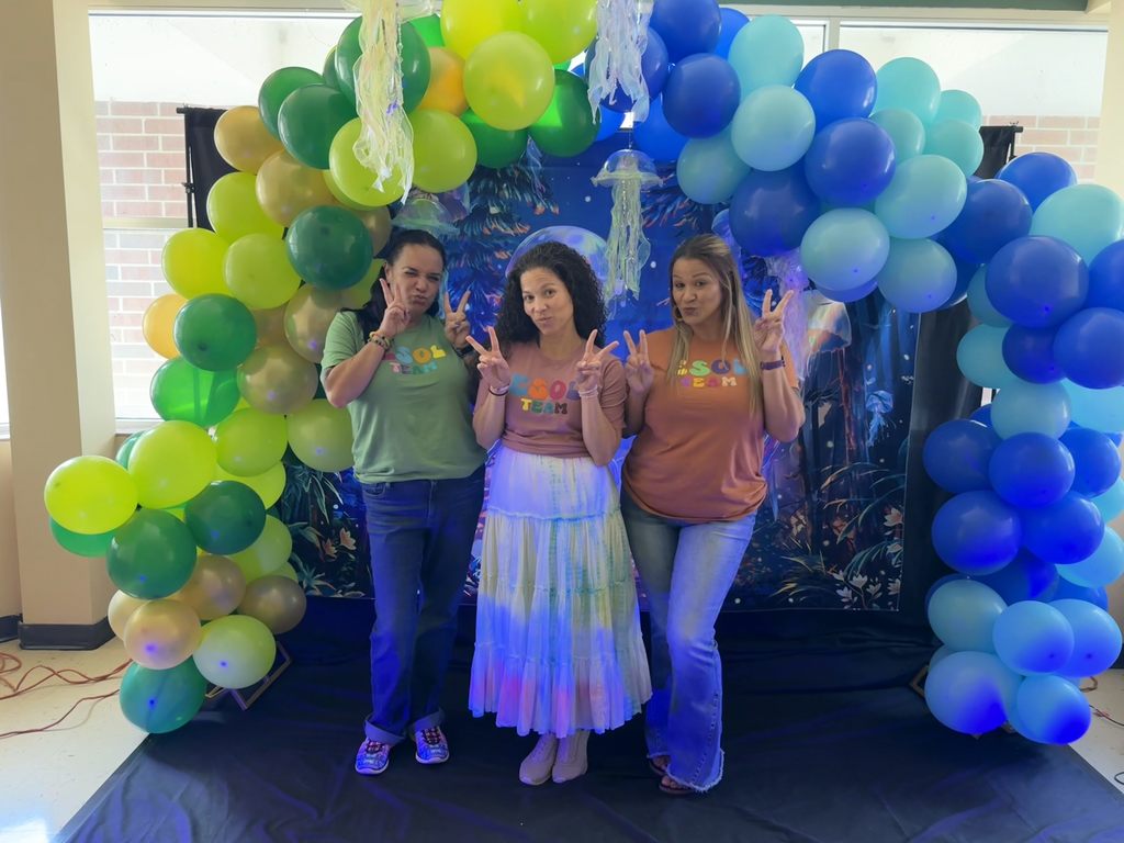 Three staff members are wearing matching "ESOL Team" shirts posing with peace signs for their photo with the balloon arch and blue light. 