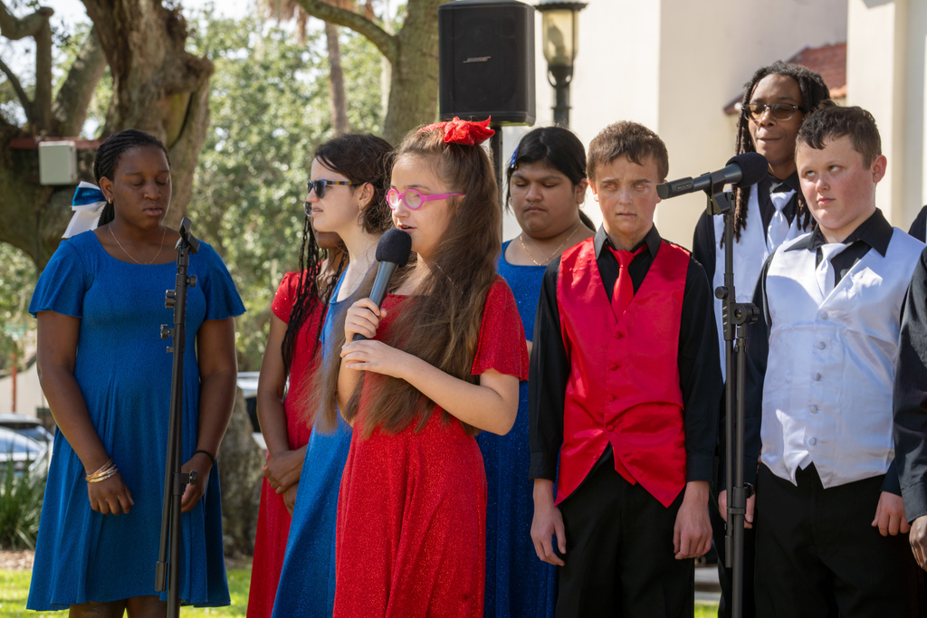 A close-up of one student wearing a red dress with red bow doing a solo outside in the plaza of downtown St. Augustine. Her classmates surround her all around. 