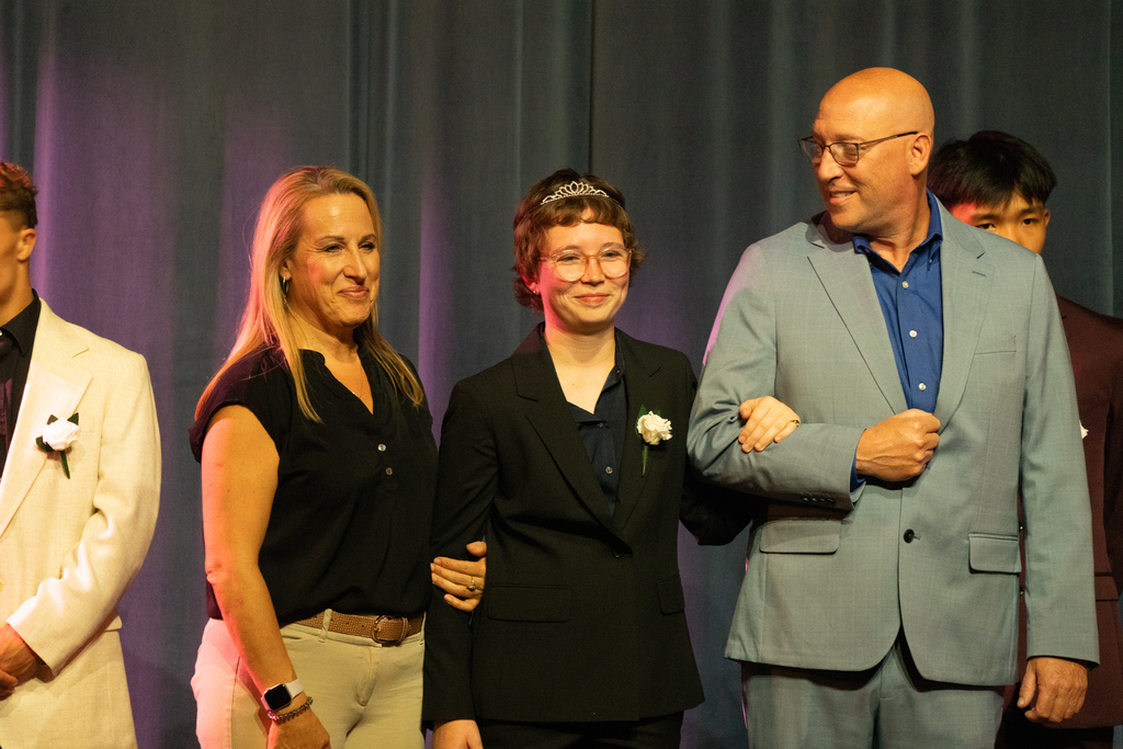 A senior is dressed in a suit holding her parents arms on stage in Kirk. 