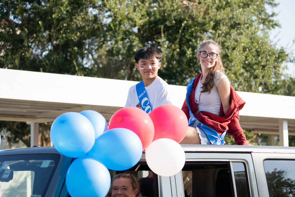Two Homecoming Court sit onto of a Jeep Wrangler with red, white and blue balloons smiling for a photo. 