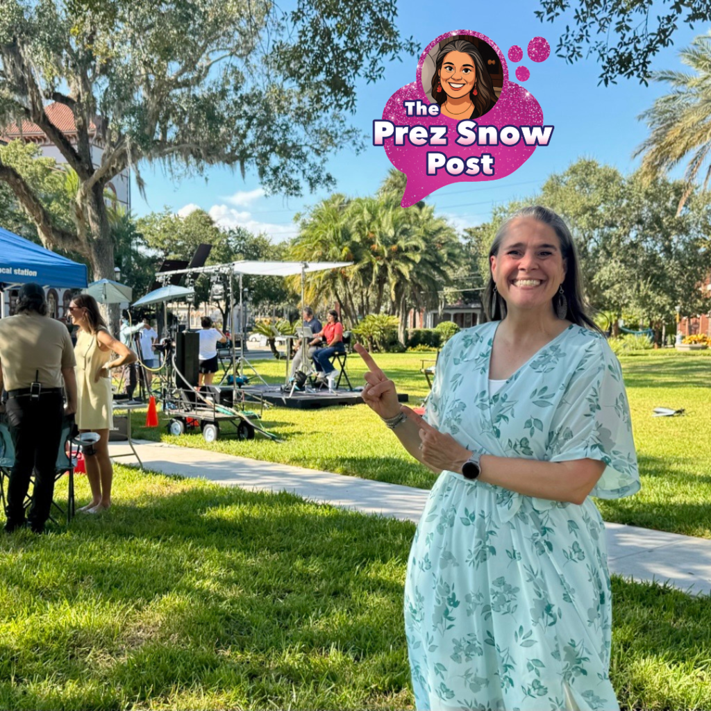 Tracie is wearing a white with green flowers scattered all over her dress pointing to the camera crew outside on Flagler College campus. 