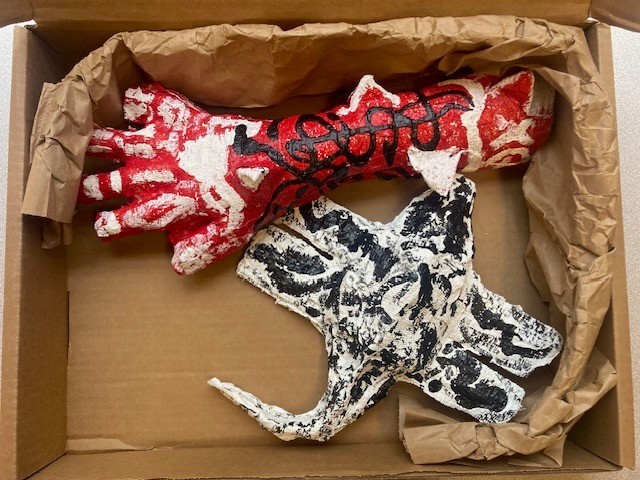 A red, white, and black plaster cloth sculpture of an arm is tucked in a brown box with a black and white mystery creature sculpture.