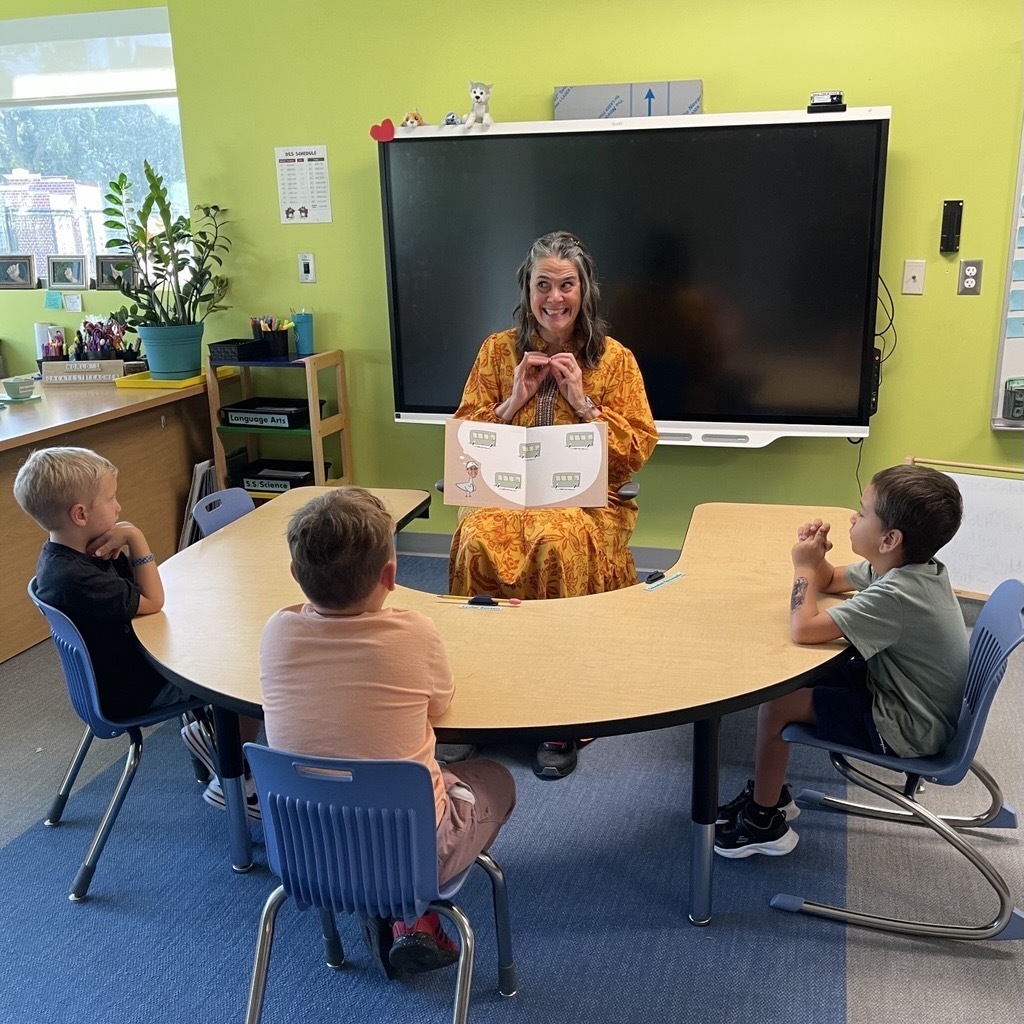 President Snow sits in a chair signing the book to the three kindergartners sitting around her. 
