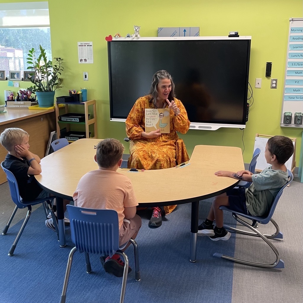 President Snow is sitting in a chair signing the book to the three kindergartners in the classroom.