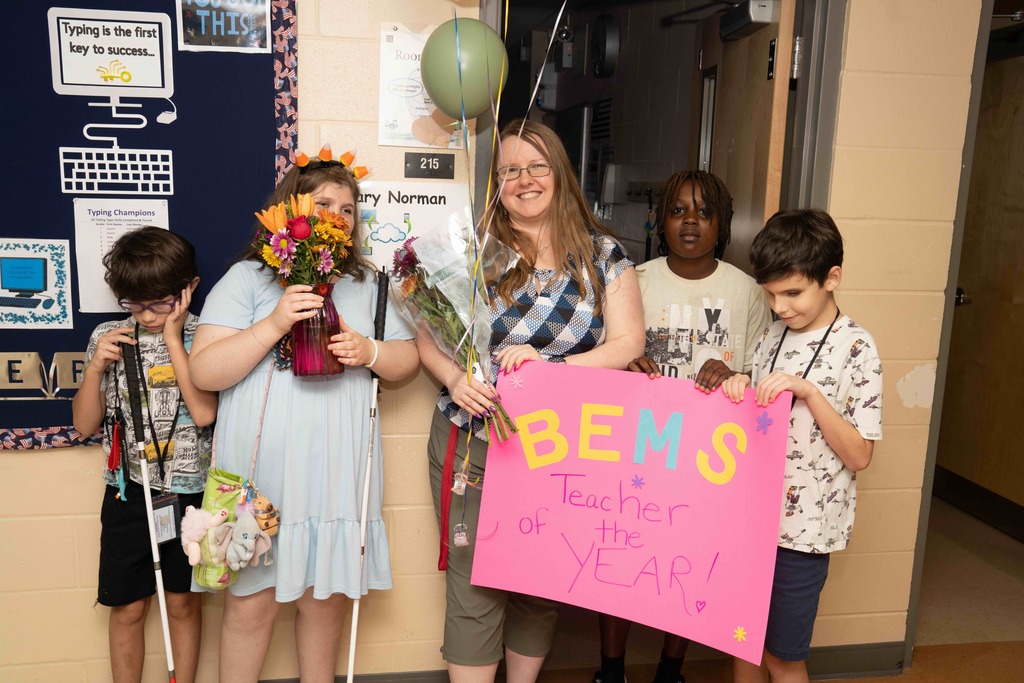 Hilary is holding "BEMS Teacher of the Year" pink sign with flowers and balloons smiling with some of her students in the hallway.