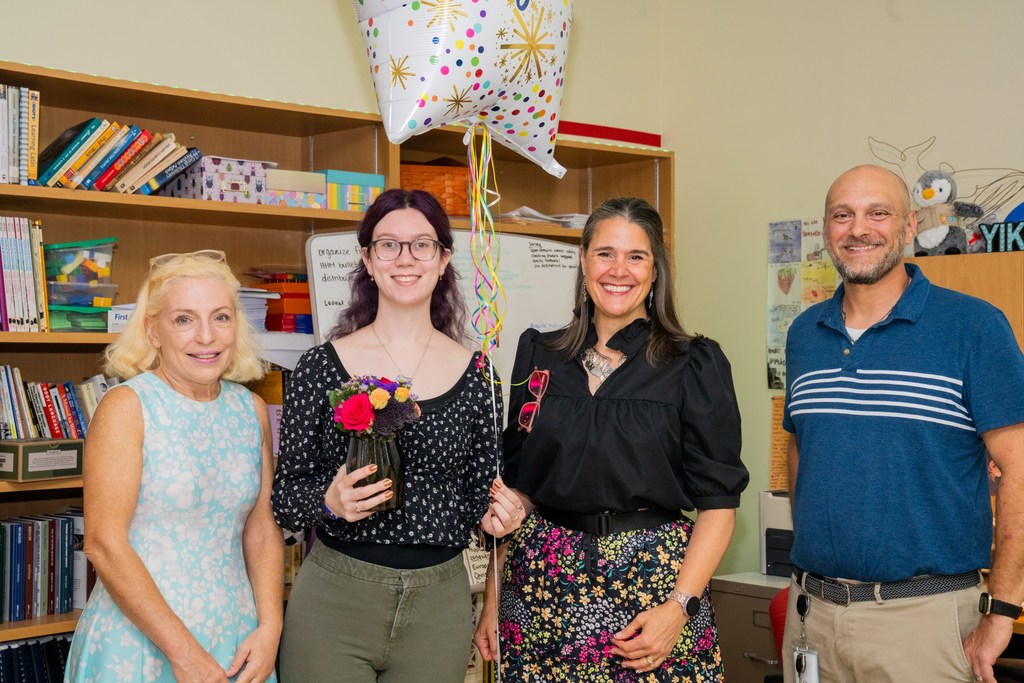 Hannah is holding flowers and balloons smiling with the Blind Principal, FSDB President, and Assistant Principal inside her classroom.