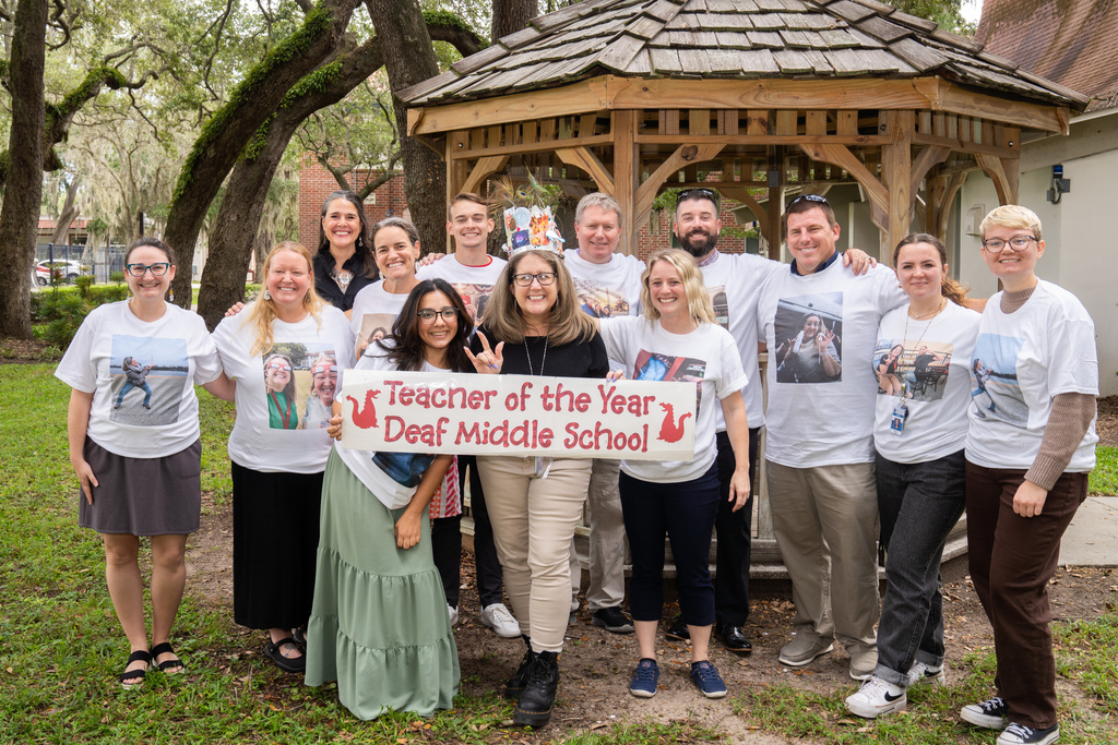 Deaf Middle School staff all wearing different white t-shirts with Nicole's pictures on them, gathered in front of the gazebo with the Teacher of the Year banner.