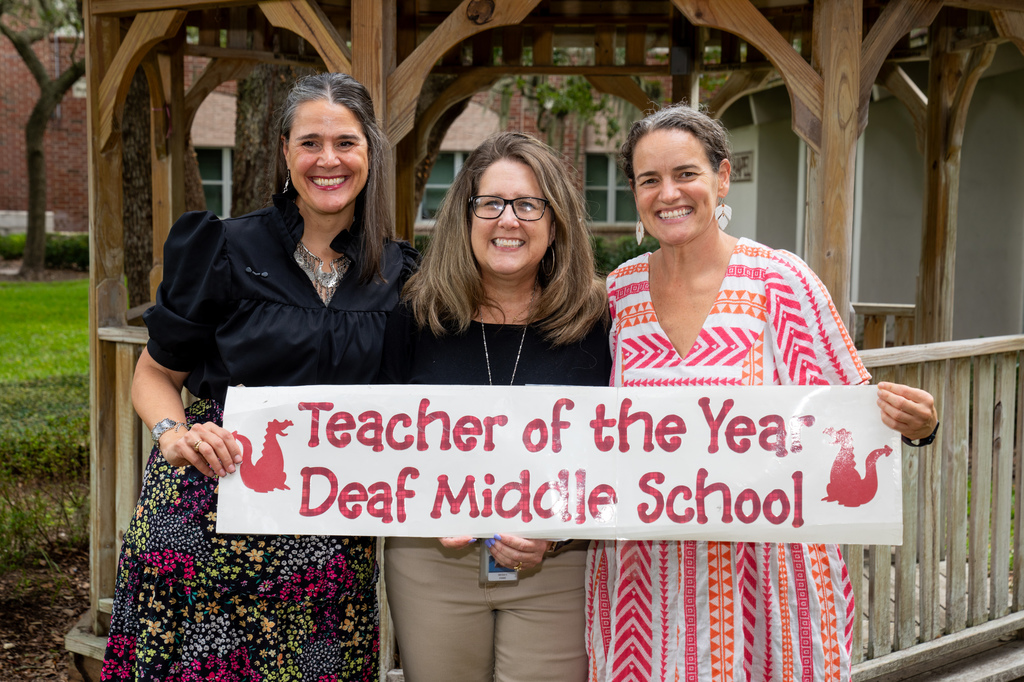 President Snow, Nicole and DMS Assistant Principal stand in front of a gazebo outside holding the Teacher of the Year banner.