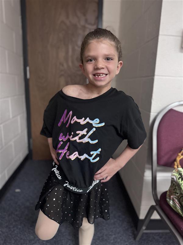 A elementary student wearing a black shirt that says "move with heart" and a black tutu skirt smiling for her photo.