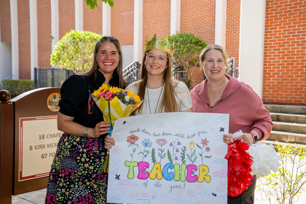 Left to Right: President Snow, Colleen, and Assistant Principal Marcelle holding a white "Teacher of the Year" sign and flowers outside of Kirk.