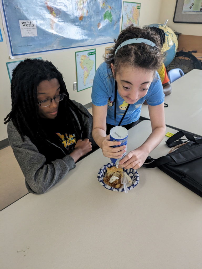 Danika S is adding whipped cream from a spray can to Derrick M's pie in a paper bowl.