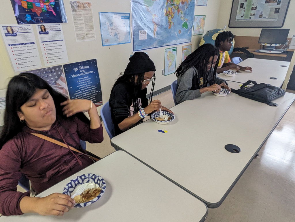 Juana, Serraya, Derrick M, and Derrick C are all seated at tables in Mr. Ward's classroom. In front of them are paper bowls and plastic forks with their pie slice in it.