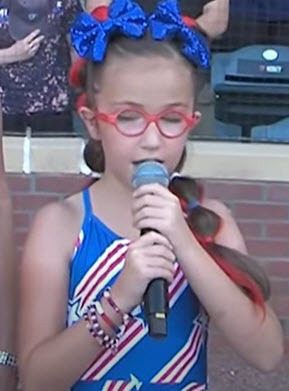 McKenzie stands holding a microphone while singing. She is wearing a blue dress with red and white stars and stripes, red, white and blue beaded bracelets, red glasses, blue bows in her hair and her long hair is in a bubble ponytail with red highlights mixed in draped over her shoulder.