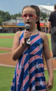 McKenzie stands on the field of the Jacksonville Jumbo Shrimp. She is wearing a blue dress with red and white stripes and stars, red glasses and her hair is double french braided. She holds a microphone while singing.
