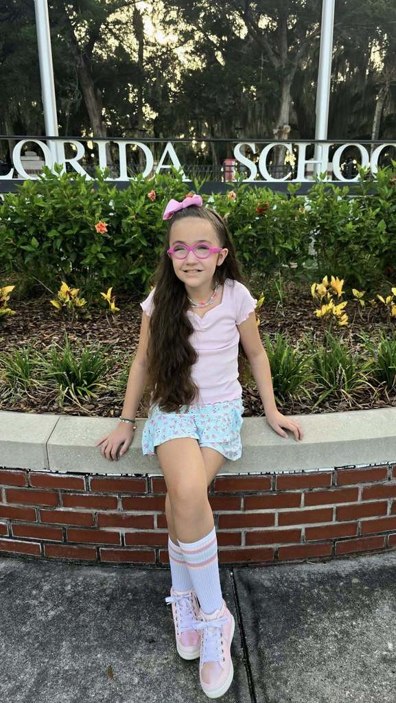 McKenzie sits on the wall in front of the FSDB entrance sign. She is wearing a blue skirt, pink top, pink glasses, pink bow, pink sneakers and white and pink socks.