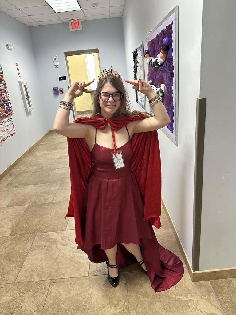 A student is wearing a red dress, red cape and a crown holding peace signs posing for a photo inside the hallway. 