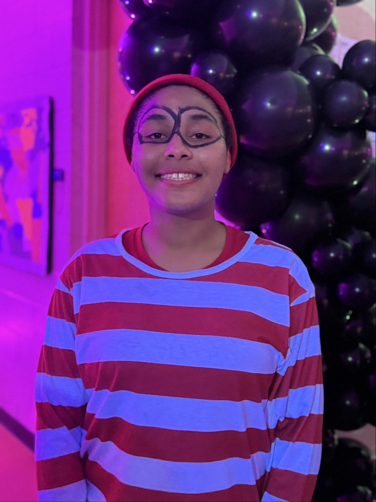 A student is dressed up like Waldo for halloween, smiles for her photo inside the dorms. 