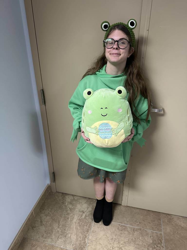 A student wearing all green and holding a frog pillow in front of her smiles for a photo in the hallway. 