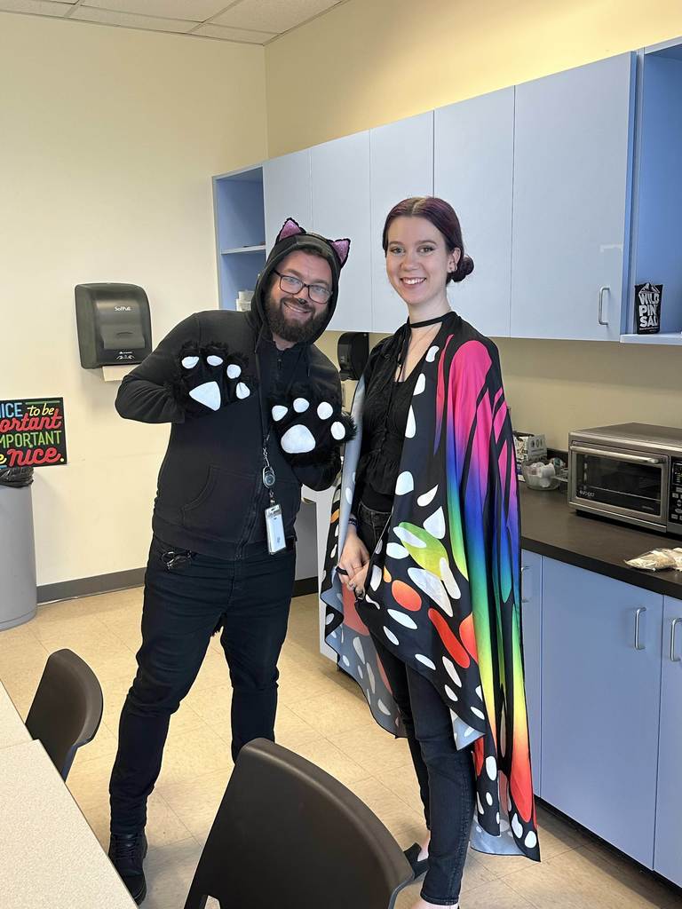 Two staff members are dressed as a cat and butterfly smiles for a photo together inside the break room. 