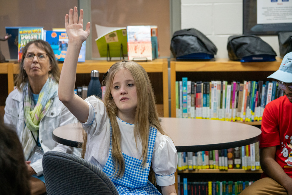 A student dressed like Dorthy is sitting at a library table and raising her hand. 