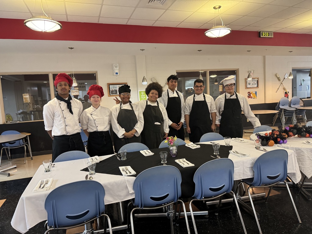 Culinary Arts students wearing red, white and black chef hats and gowns smile for a photo next to the table they set up for Teacher of the Year winners. 
