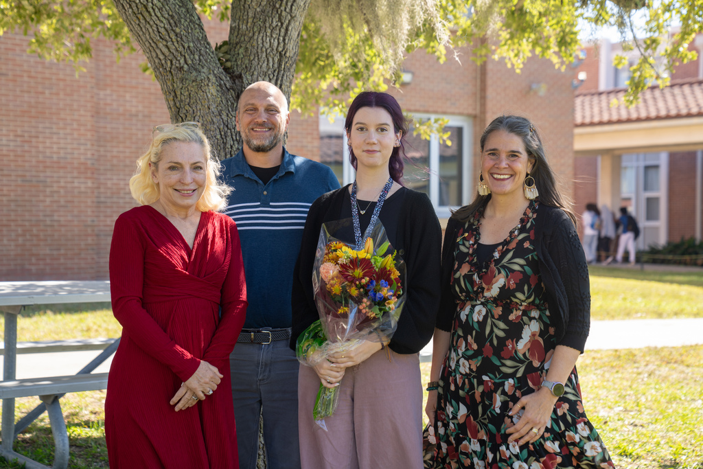 Carol Bogue (wearing a red dress with blonde hair), Justin Cosgrove (wearing a blue polo with five white stripes across the chest and gray pants), Hannah Glynn holding her flowers, and Tracie Snow (wearing a black floral dress with cream, burgundy, and green flowers with a black cardigan.  Her hair is pulled off her face and she has gold dangly earrings).