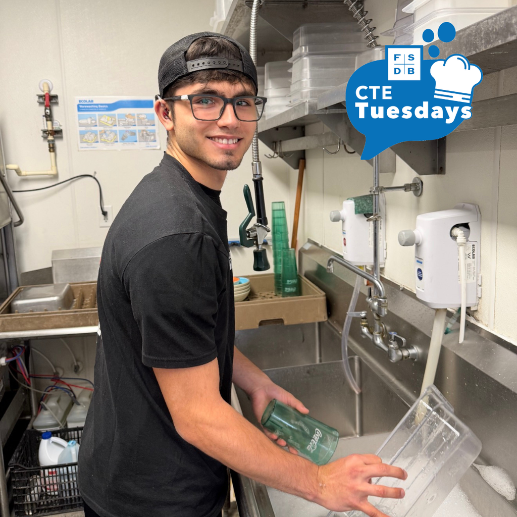 A student worker is wearing black clothing smiling for the photo while he is washing dishes inside a restaurant's kitchen.