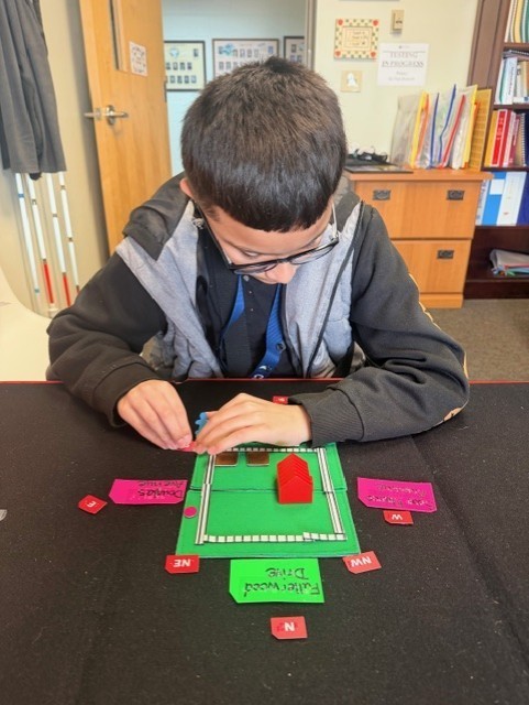 Manny looks down and works on his tactile map inside the office. 
