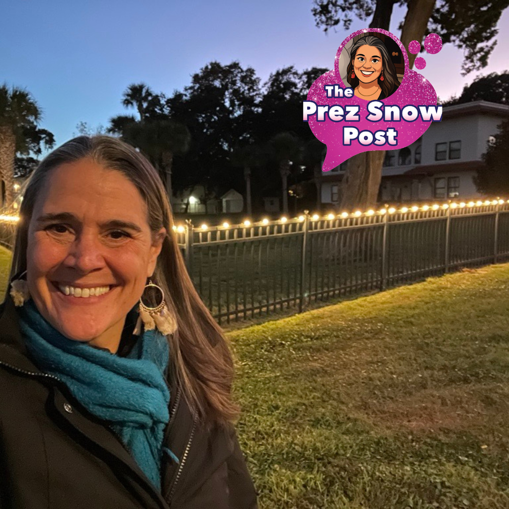 Tracie is wearing a blue scarf and black jacket smiling for a selfie with the lights on her fence of the President's House.