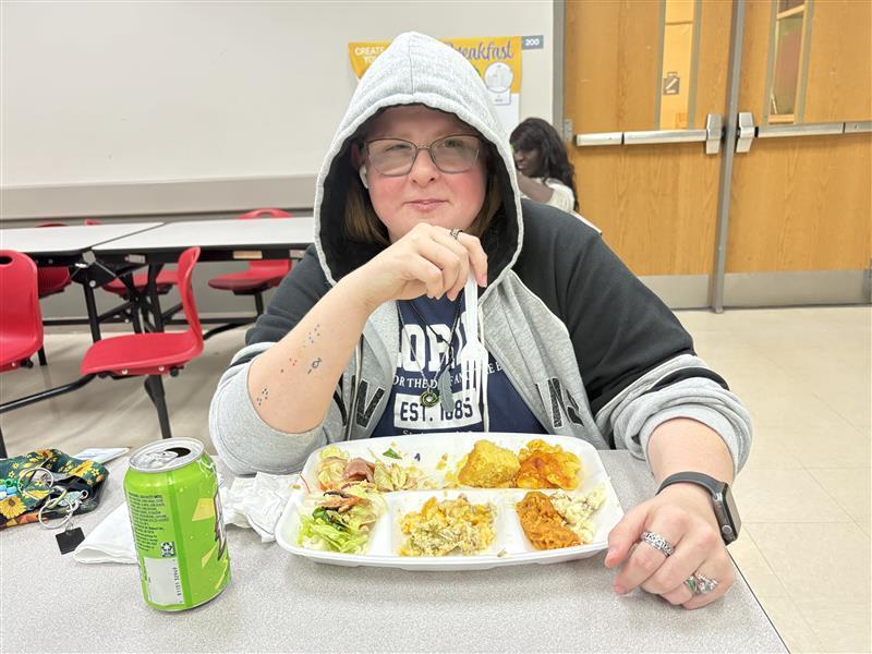A student is smiling with her Thanksgiving meal sitting down at a table inside the cafeteria.