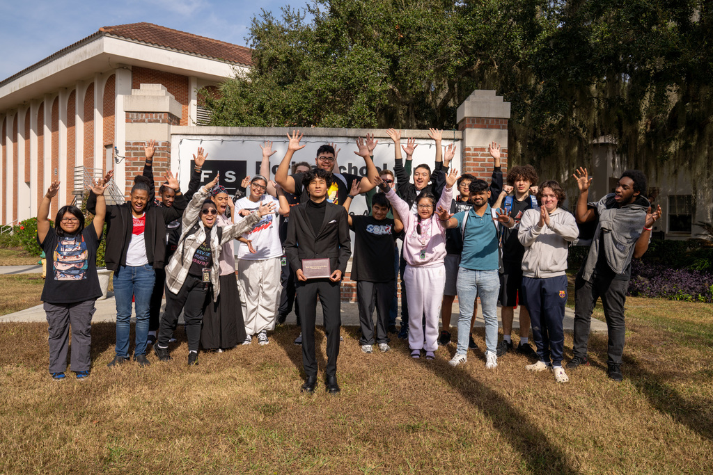 Clark stands in the middle holding his diploma while his peers gather around him with hands in the air by the FSDB sign.