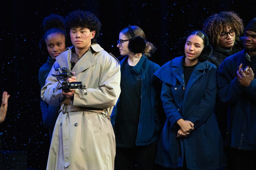 A camera man wearing a tan jacket and holding the video camera on stage in Kirk.
