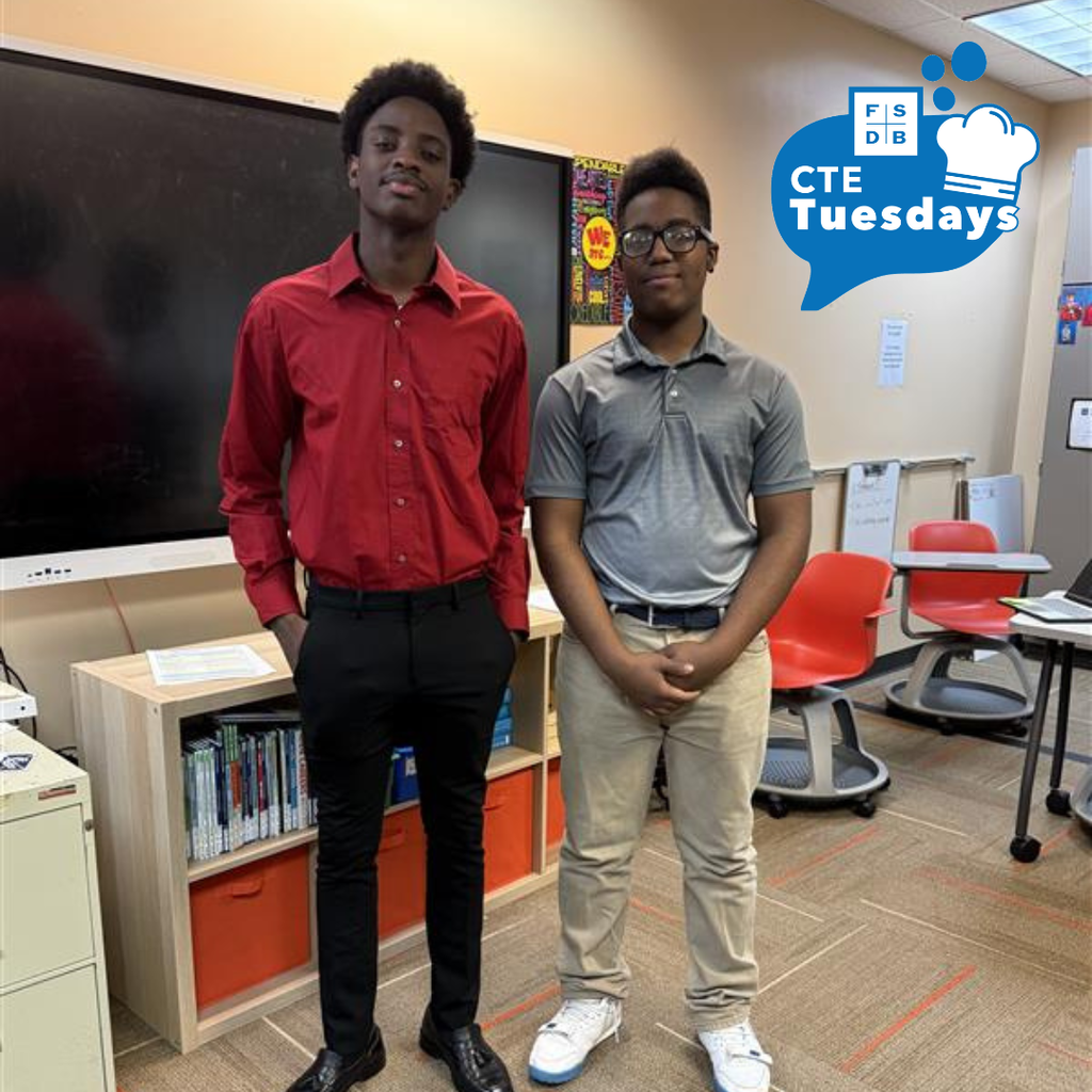 Two students dressed up in nice shirts and pants smile for a photo inside a classroom.