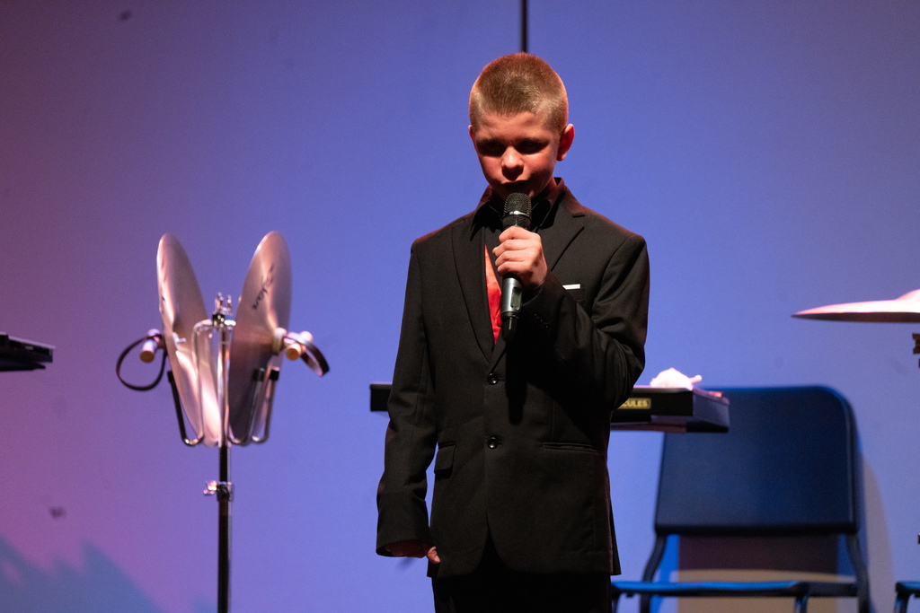 A blind student wearing a black suit holding a microphone singing on stage in the Music building.