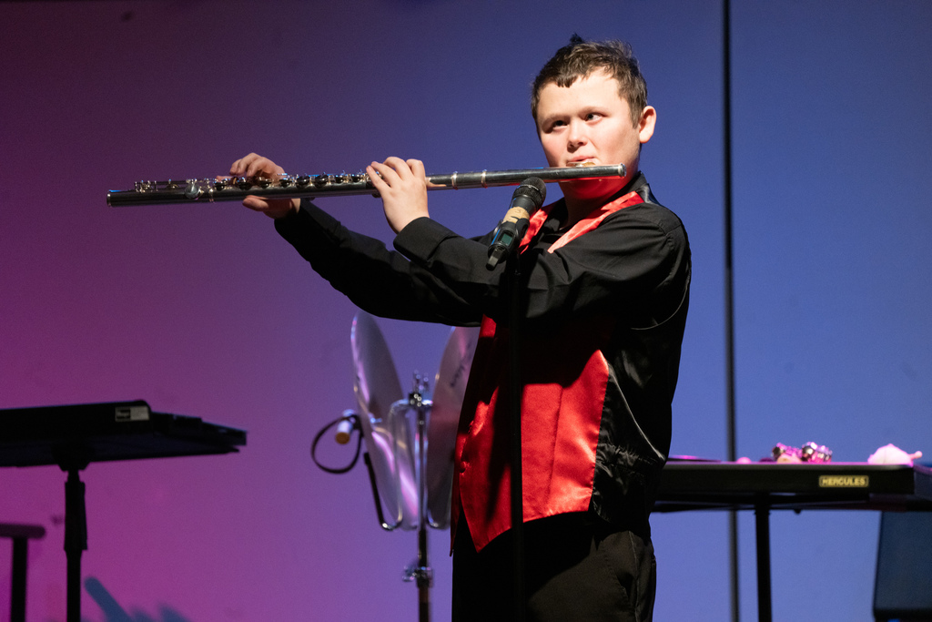 A blind student wearing a black and red suit play the flute on stage in the Music building.
