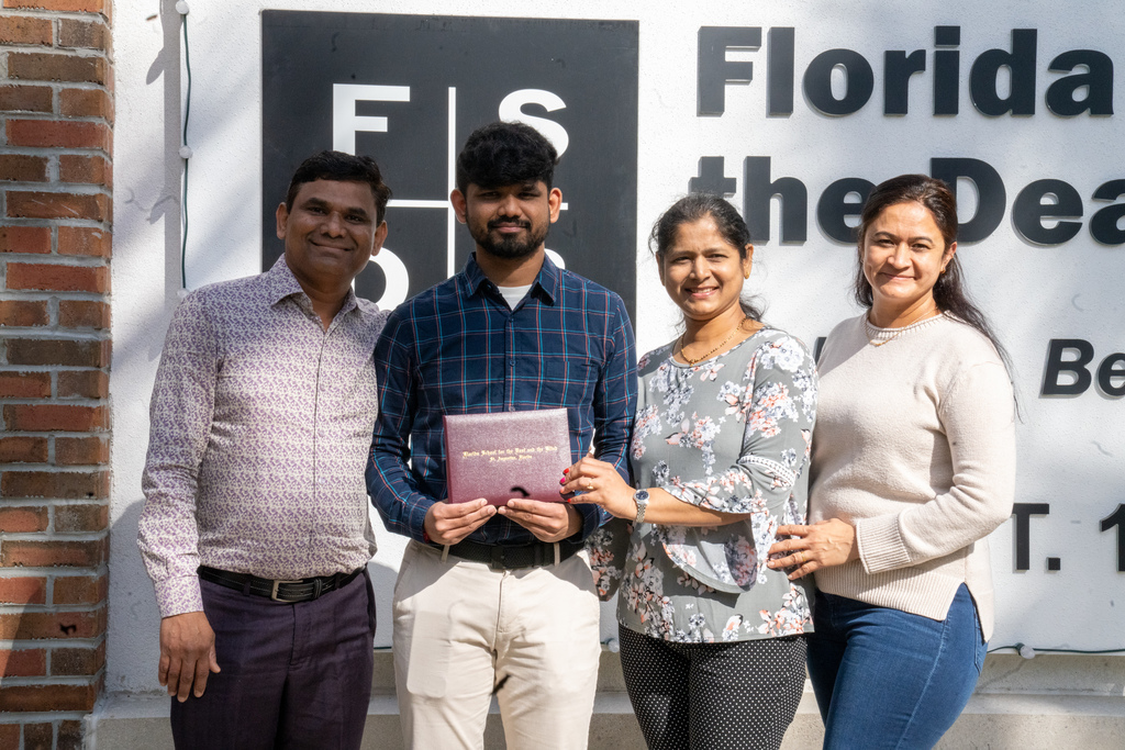Sujal holding his diploma and smiling with his family by the FSDB sign outside. 