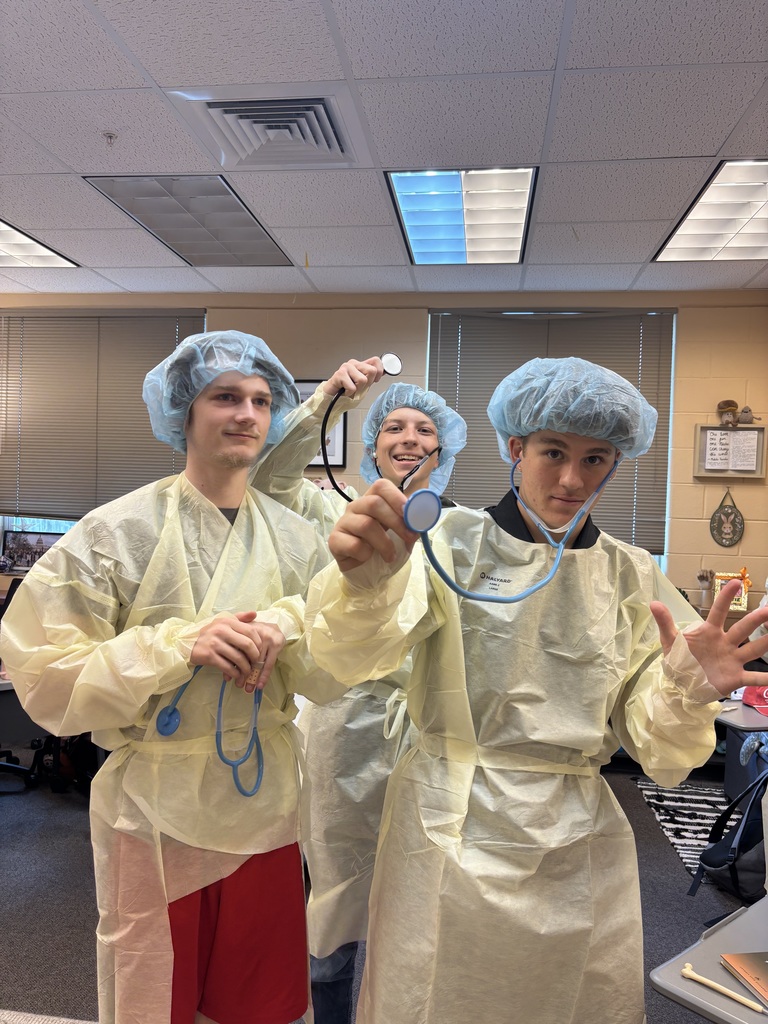 Three students dressed in PPE and holding stethoscopes for a photo inside the classroom.
