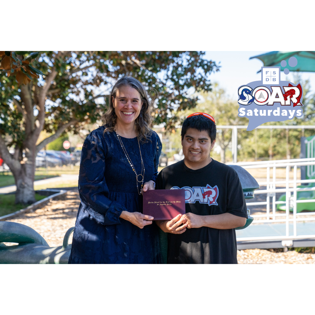 President Snow wearing a blue dress smiling with Max who is holding his diploma outside of the Kramer playground.