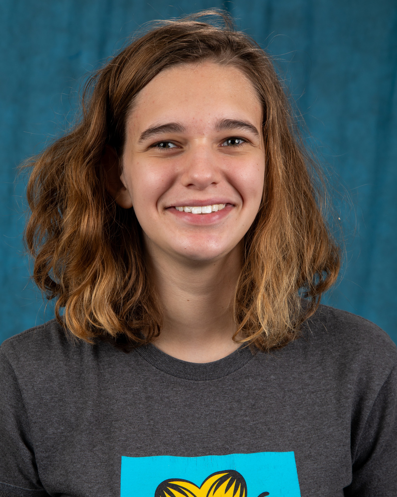 7th grade photo of Sophia. She has wavy brunette hair that is shoulder length and smiling at the camera. She is wearing a grey t-shirt and standing in front of a blue background.