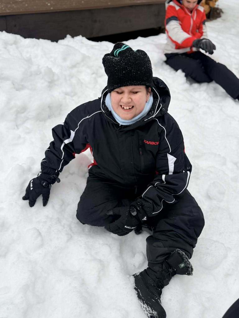 A blind student wearing black hat, jacket and pants smiles sitting in the snow. 