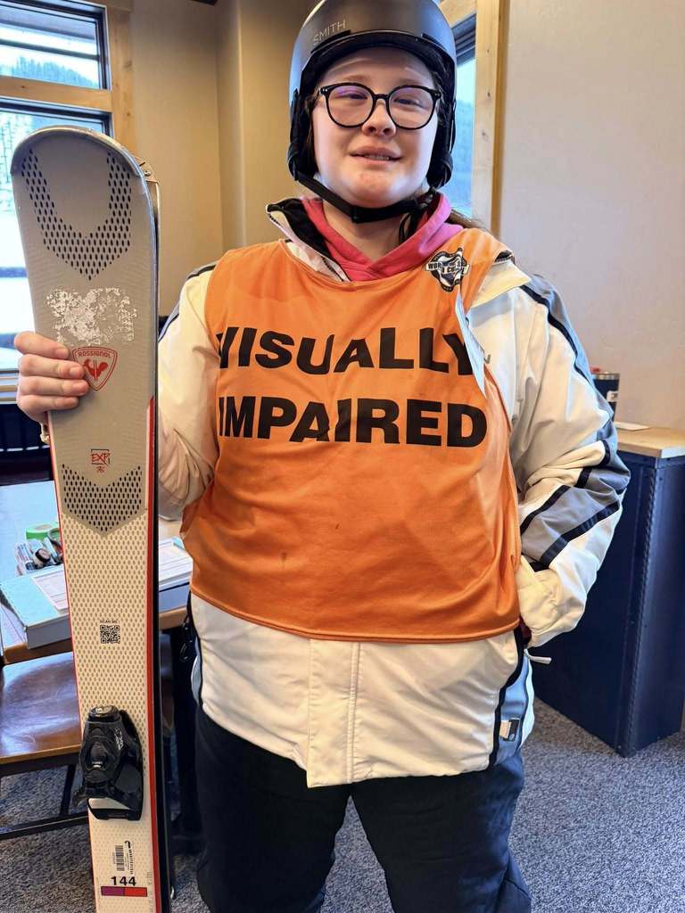A blind student wears her "visually impaired" vest smiling with her skis inside the building. 