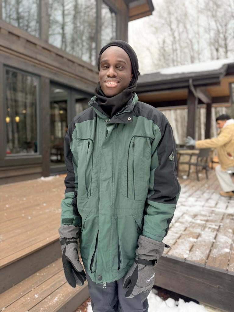 A blind student wearing a green jacket and black winter hat smiles for his photo outside their cabin. 