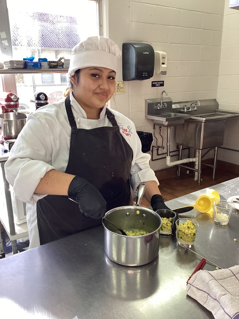 Ashley smiles wearing a white chef apron and hat, working on her dish of Elote corn for the skills completion. 