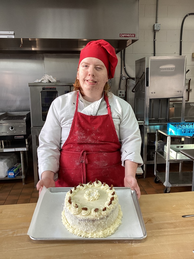 Jacob wearing a white chef jacket with red apron and hat smiles with his cake he practiced for the baking competition. 