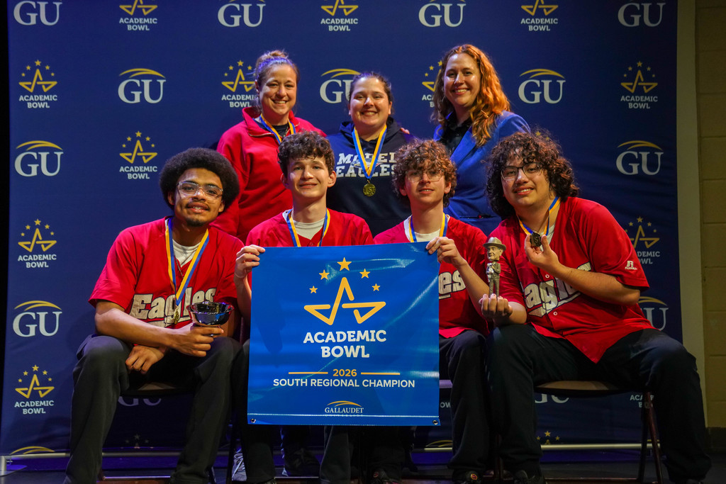 MSSD academic bowl group smile for a photo with their medals and banner that says "Academic Bowl South Regional Champion" on stage in Kirk.