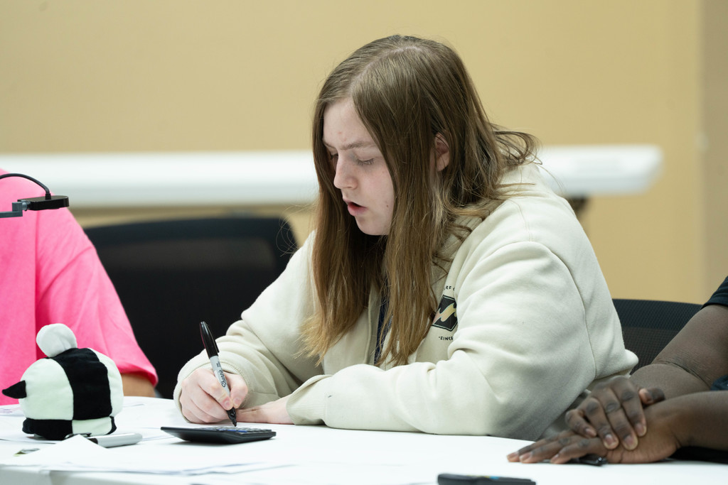 A student writing her answer on a piece of paper inside a classroom.