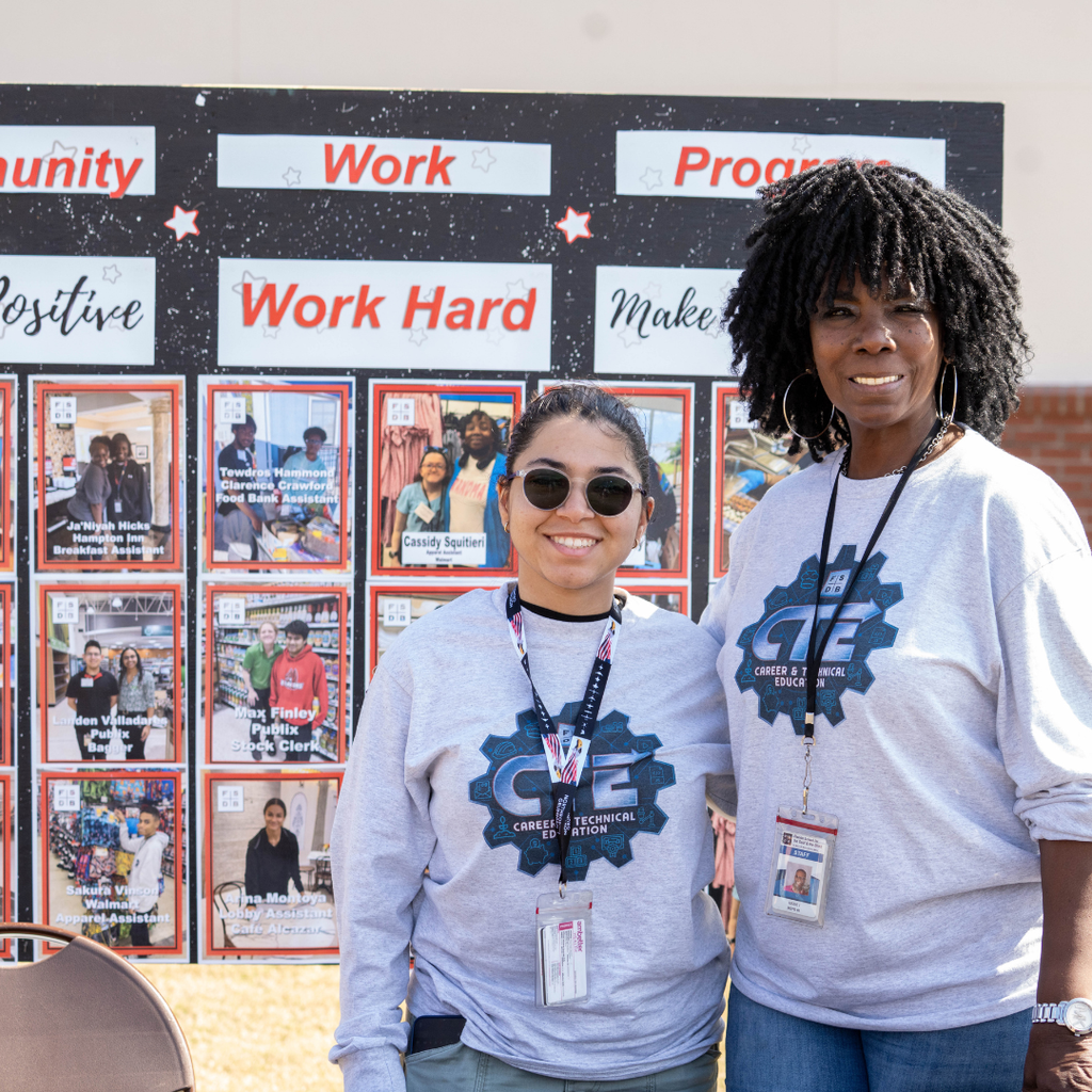 Staff member smiles with a SOAR student by the "Community Work Program" poster board outside.